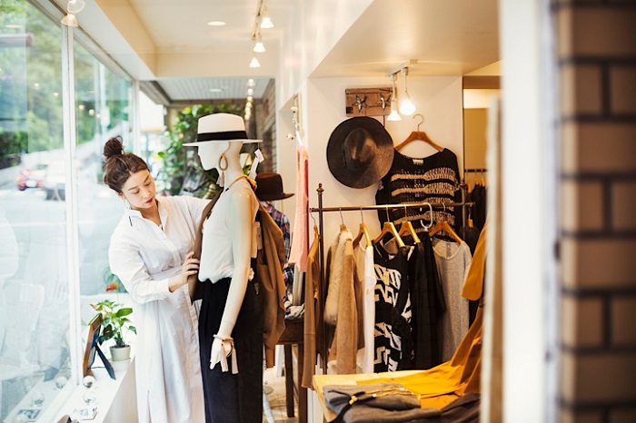 Shop assistant adjusts a mannequin in a bright boutique with racks of clothing and hats on display.