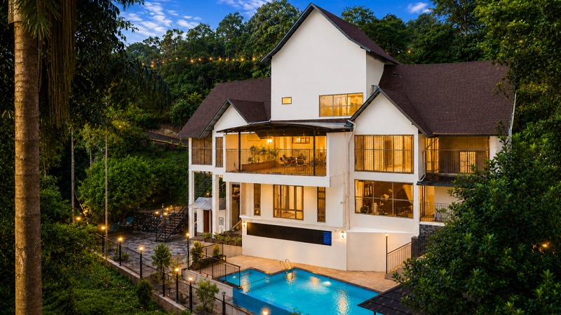 White multi-story villa with a blue rectangular pool, string lights, and surrounding trees at dusk.