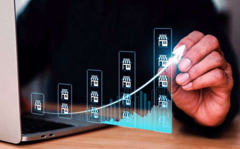 Hand drawing an upward-trending chart made of storefront icons beside a laptop.