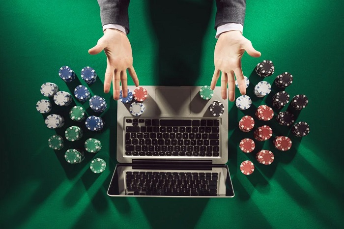 Overhead view of a laptop on a green casino table, hands placing poker chips around the keyboard for online gambling.