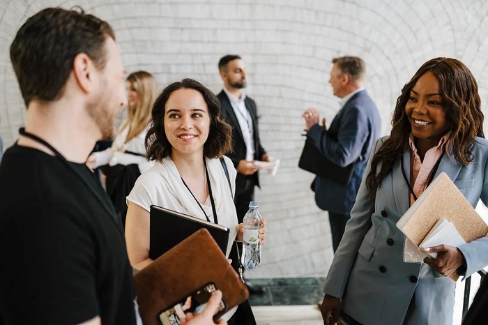 Professional networking scene in a modern hallway: a smiling woman in white holds a water bottle and folder while chatting with a man, with others conversing in the background.
